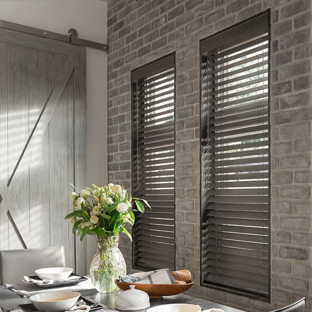 Natural wood blinds in dark gray within two tall windows side by side on a gray brick wall. Inside a dining room.