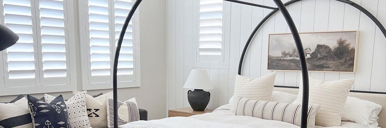 White and beige style bedroom with white Polywood shutters on a window next to a large bed.