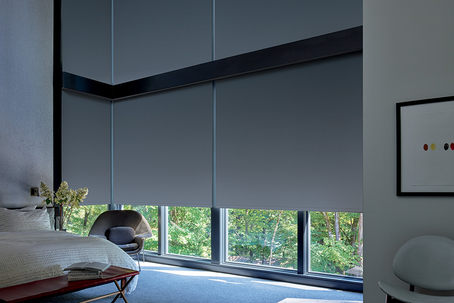 Dark gray blackout roller shades on a large wall window next to a beige style bed.