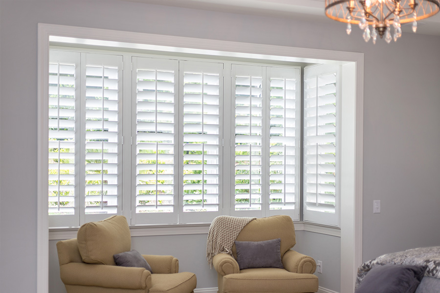 A large window with white Polywood shutters inside a bedroom nook with seating.