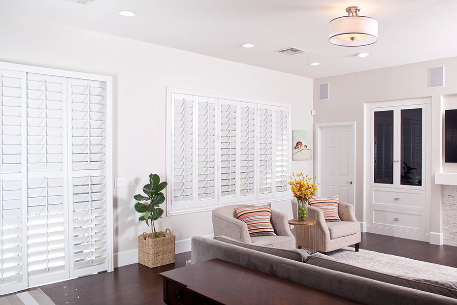 Known by many names, plantation shutters offer exceptional style, energy efficiency, durability, and light control. White Polywood shutters covering a window and sliding door in a living room
