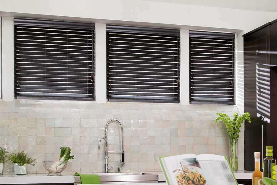 When deciding between wood blinds vs. faux wood blinds, pick moisture-resistant faux wood for the kitchen. Large modern kitchen with four small square-sized windows displaying dark wood blinds.