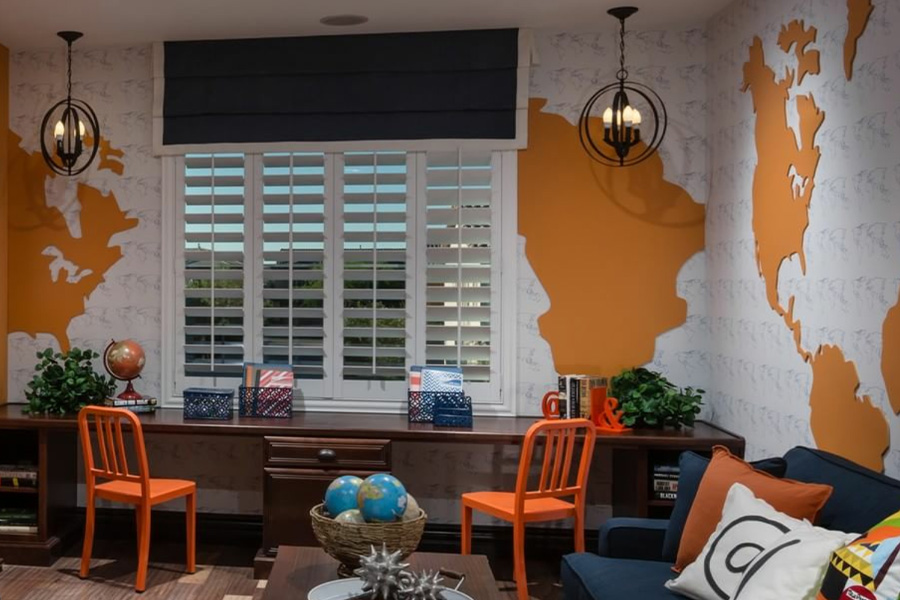 Study room with orange world map wall design, white plantation shutters, orange chairs at wooden desk, pendant lights, globes on coffee table, and navy couch with decorative pillows.