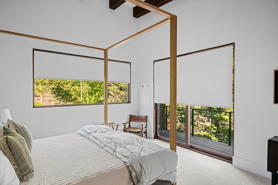 Schedule motorized shades to lower at night and open in the morning to welcome in beautiful natural light. White, motorized shades covering a sliding door and large window in a bedroom