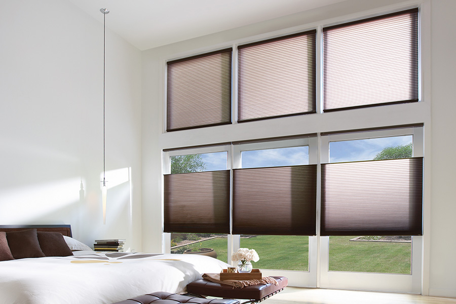 Cellular window shades keep your bedroom at a stable and comfortable temperature. rown cellular shades on a tall wall of windows within a bedroom.
