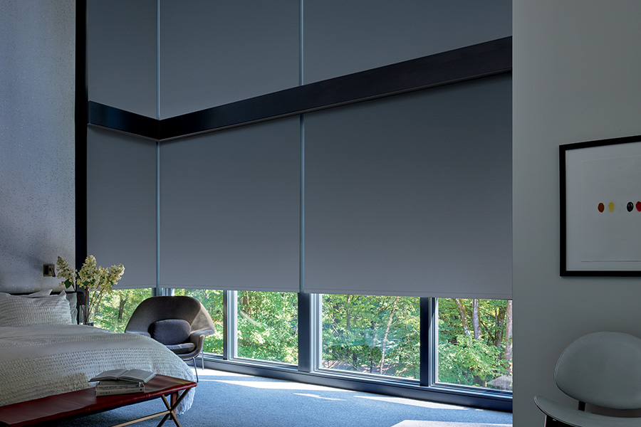 Choose blackout roller shades to keep your bedroom dark and cool. Dark gray blackout roller shades on a large wall window next to a beige style bed.