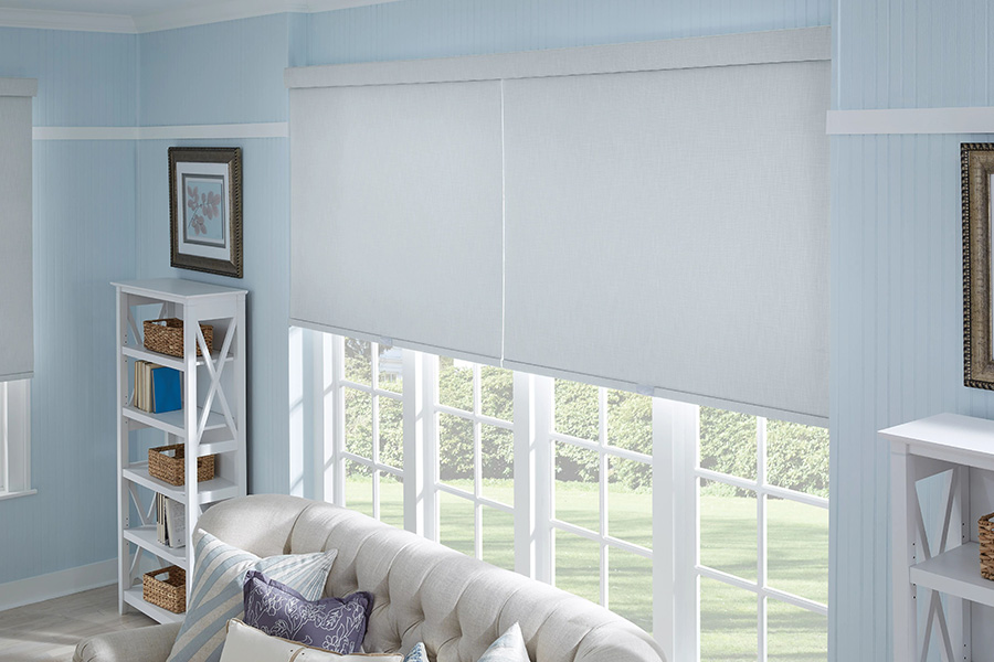 Find it hard to block drafts on a large or hard-to-reach window? Automated window shades make it easy to cover a cold window with the push of a button. Large white window shades on tall windows behind a sofa