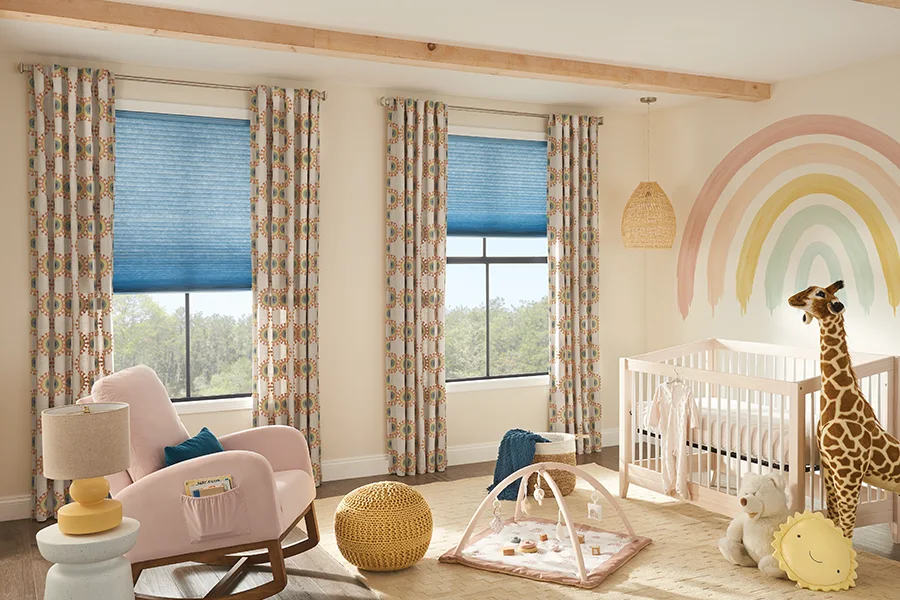 Motorization lets you adjust the shades in a nursery without disturbing your baby. slate blue cellular shades on two tall windows within a beige and pastel color nursery