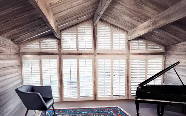 Angled white shutter window treatment on rake type windows in a lounge room area with a piano and a relaxing black chair