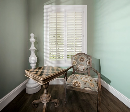 white polywood shutters on a tall window within a small nook with a table and chair plus a chess decor piece