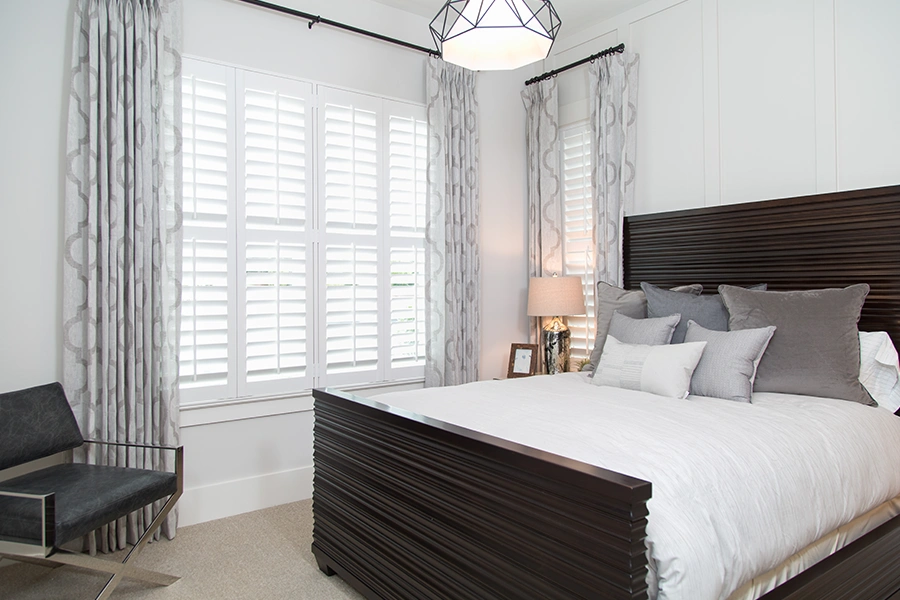 Window treatments like interior shutters and drapery help keep your home at a comfortable temperature. Gray patterned drapes and white Polywood shutters on bedroom windows