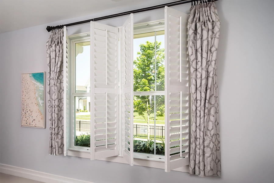Layer energy-efficient window treatments for the ultimate in style and protection. Gray patterned drapes paired with open white Polywood shutters on a window